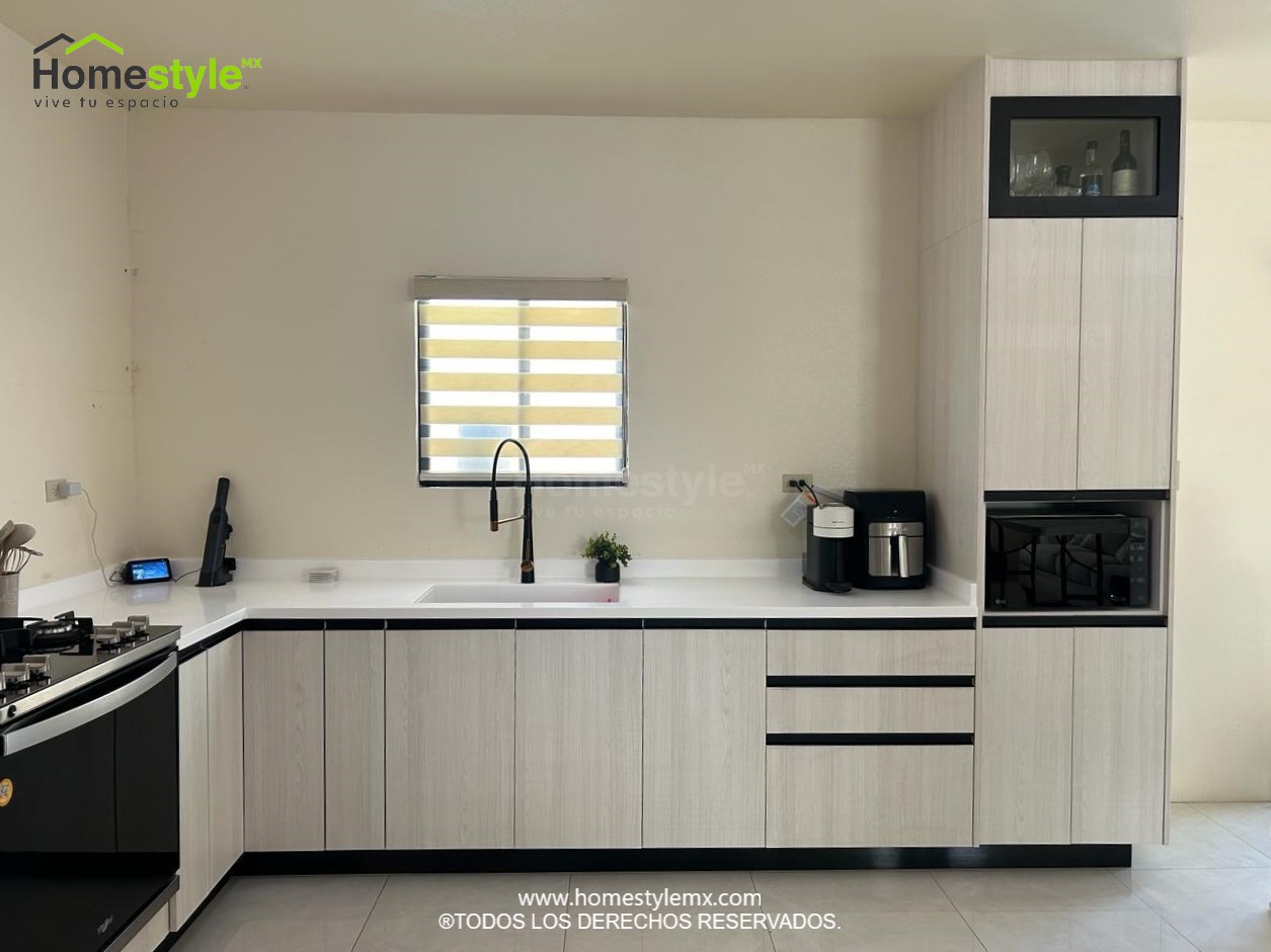 Cocina en forma de L. Diseñada en Melamina Teka Ártico para toda la gabinetería, con detalles en Melamina Negra. Con una cubierta de Superficie Solida Frosty White.